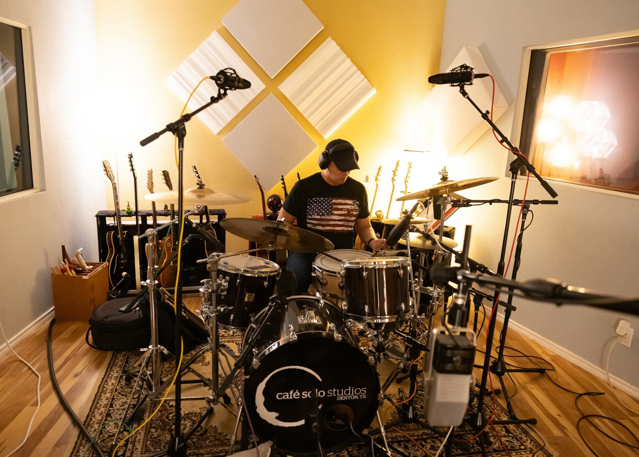 Person sitting at drum set in recording studio with microphones and soundproofing panels.