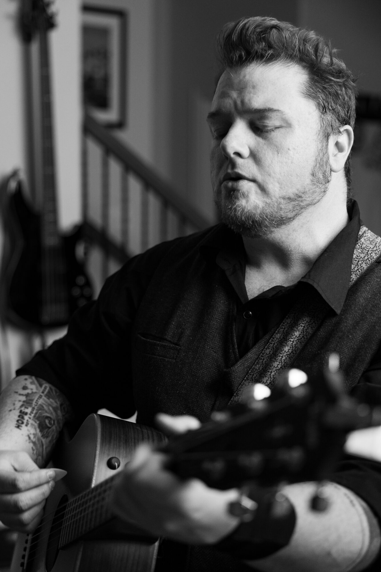 Man with beard playing guitar indoors, eyes closed, focused expression, black shirt, tattoos on arms, staircase in background.