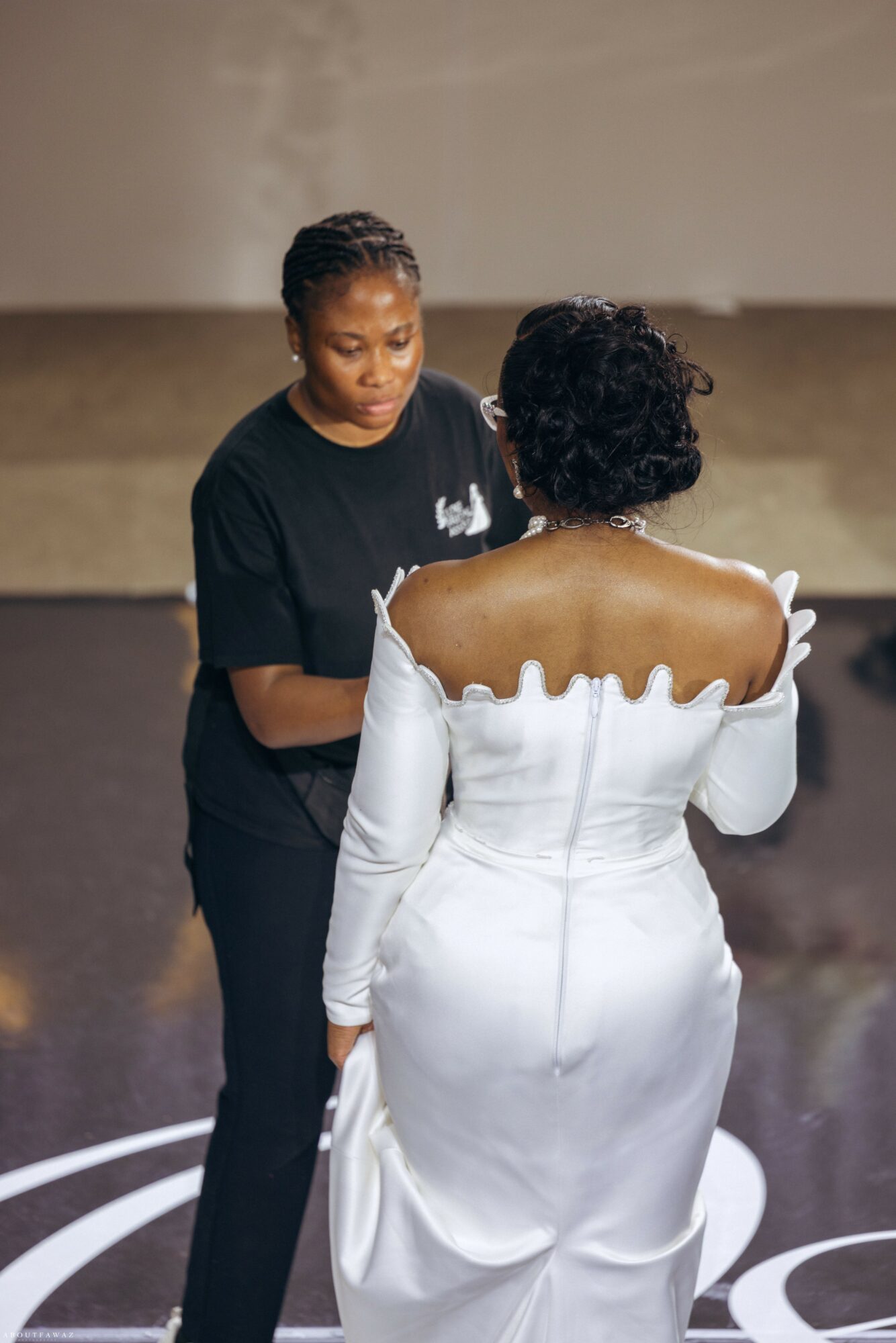 Two women stand facing each other, one in a black shirt and the other in a white dress, indoors.