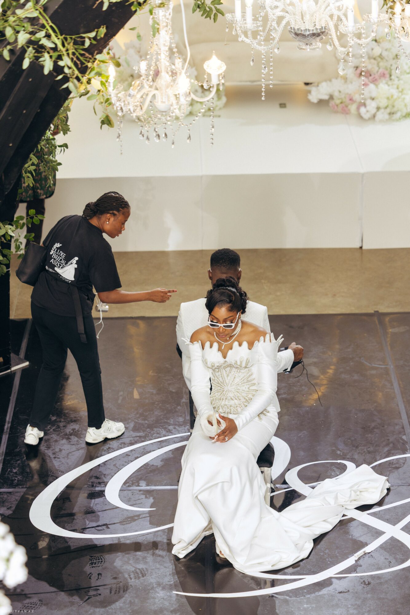 Two women in white dresses sitting on a stage with a dark floor and floral decorations above, one holding a microphone.