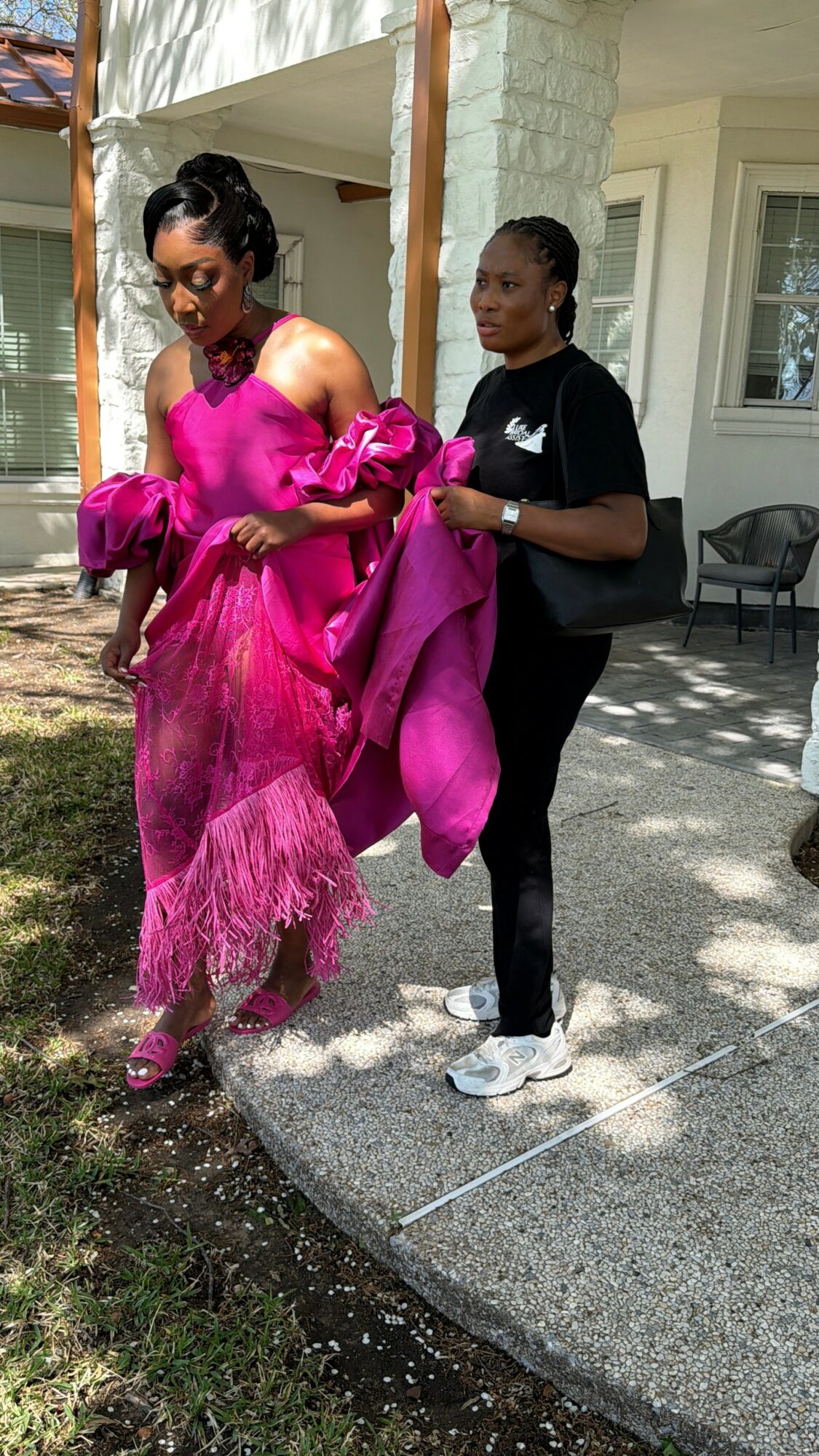 Two women standing outside a house, one in a bright pink dress, the other in black, holding a pink garment.