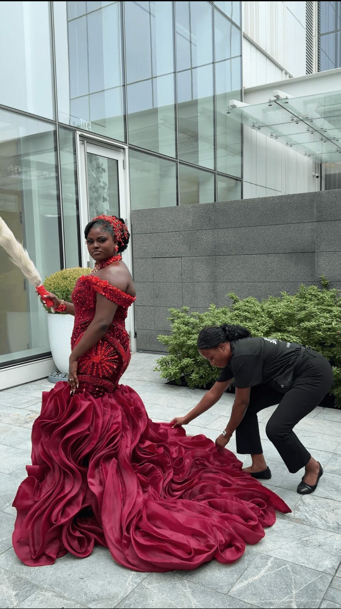 Woman in a red gown with a large train, standing outside a modern glass building, with a person adjusting the dress.