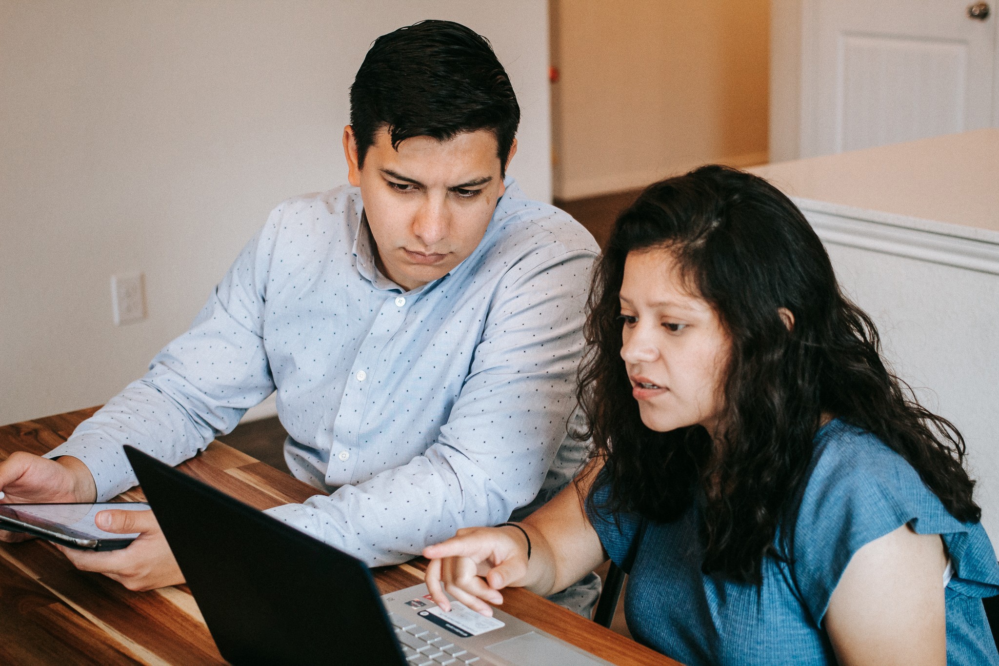 Two people sitting at a table, looking at a laptop and a tablet, with a neutral background.