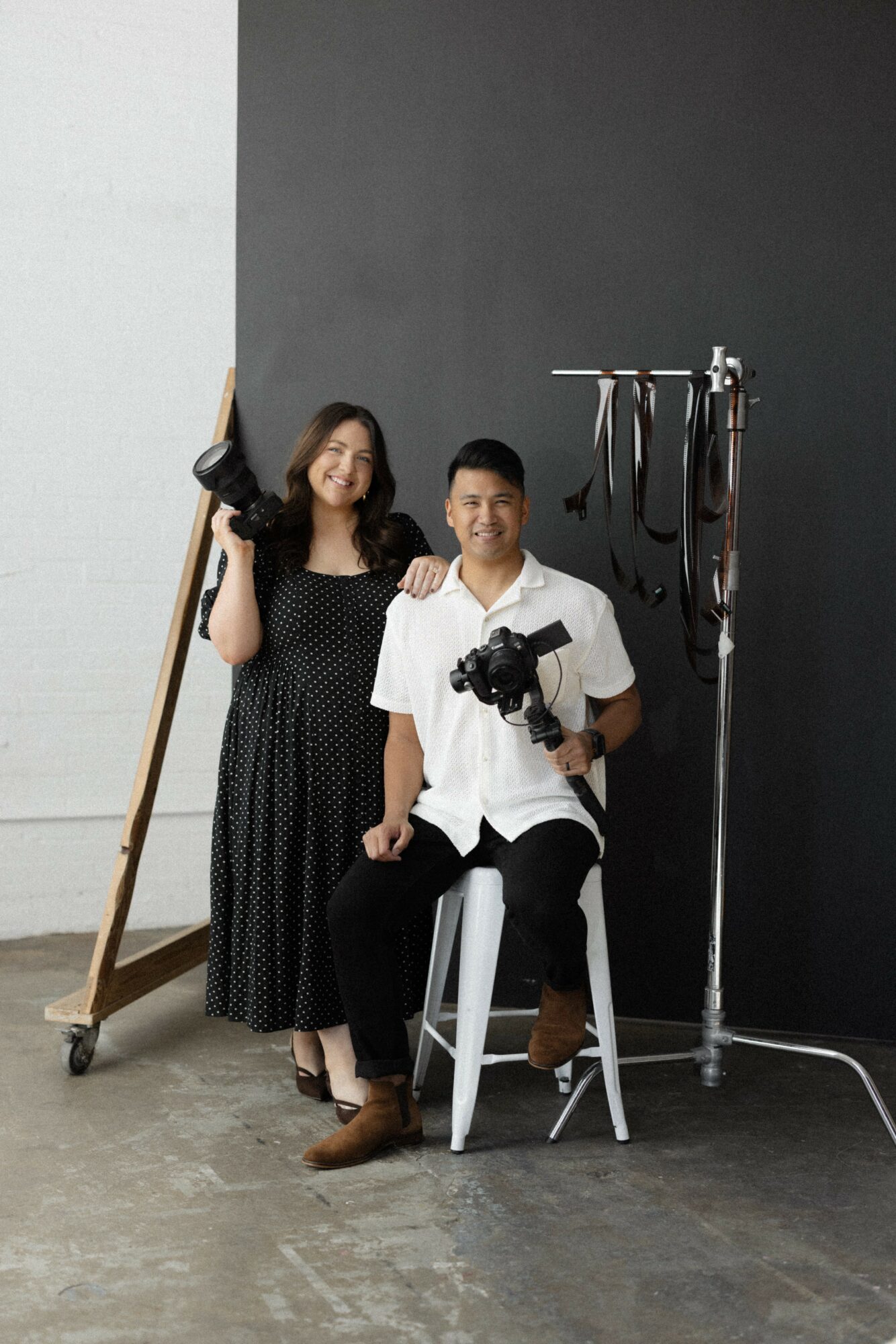 Two people, one seated and one standing, in a photography studio with dark and light walls, holding equipment.