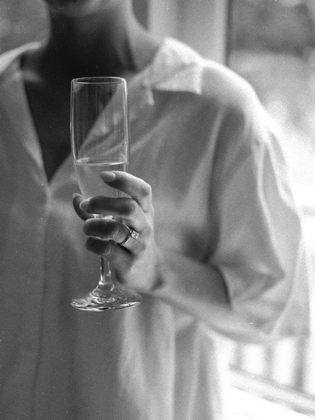 Person holding a champagne flute with a blurred background, wearing a light-colored shirt, in black and white.