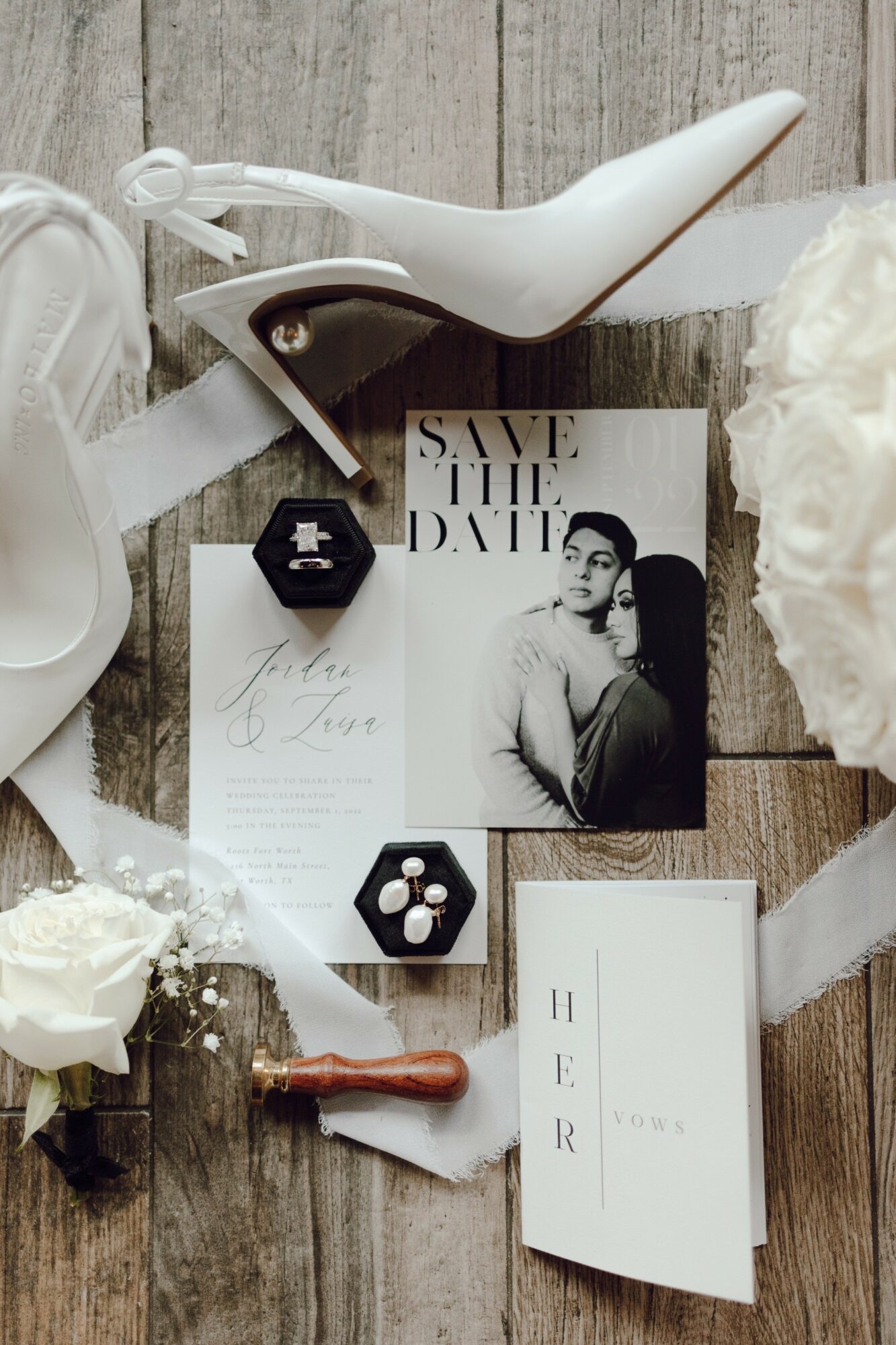 Wedding invitation with black and white photo, white roses, bird figurine, and small hexagonal boxes on wooden surface.