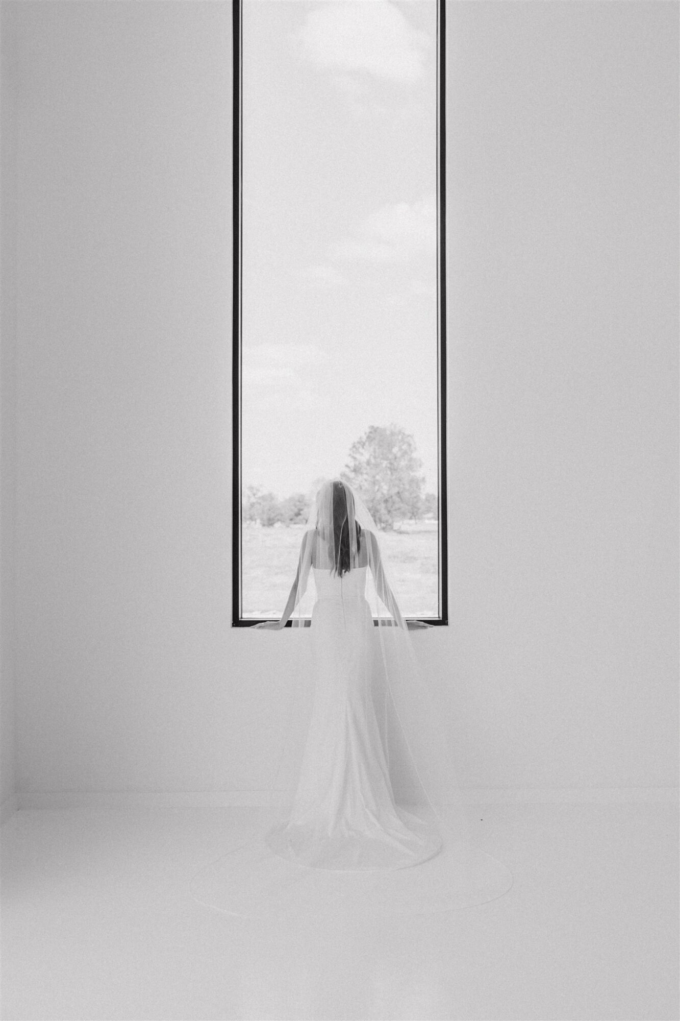 Person in a white dress standing in front of a tall window with a view outside.