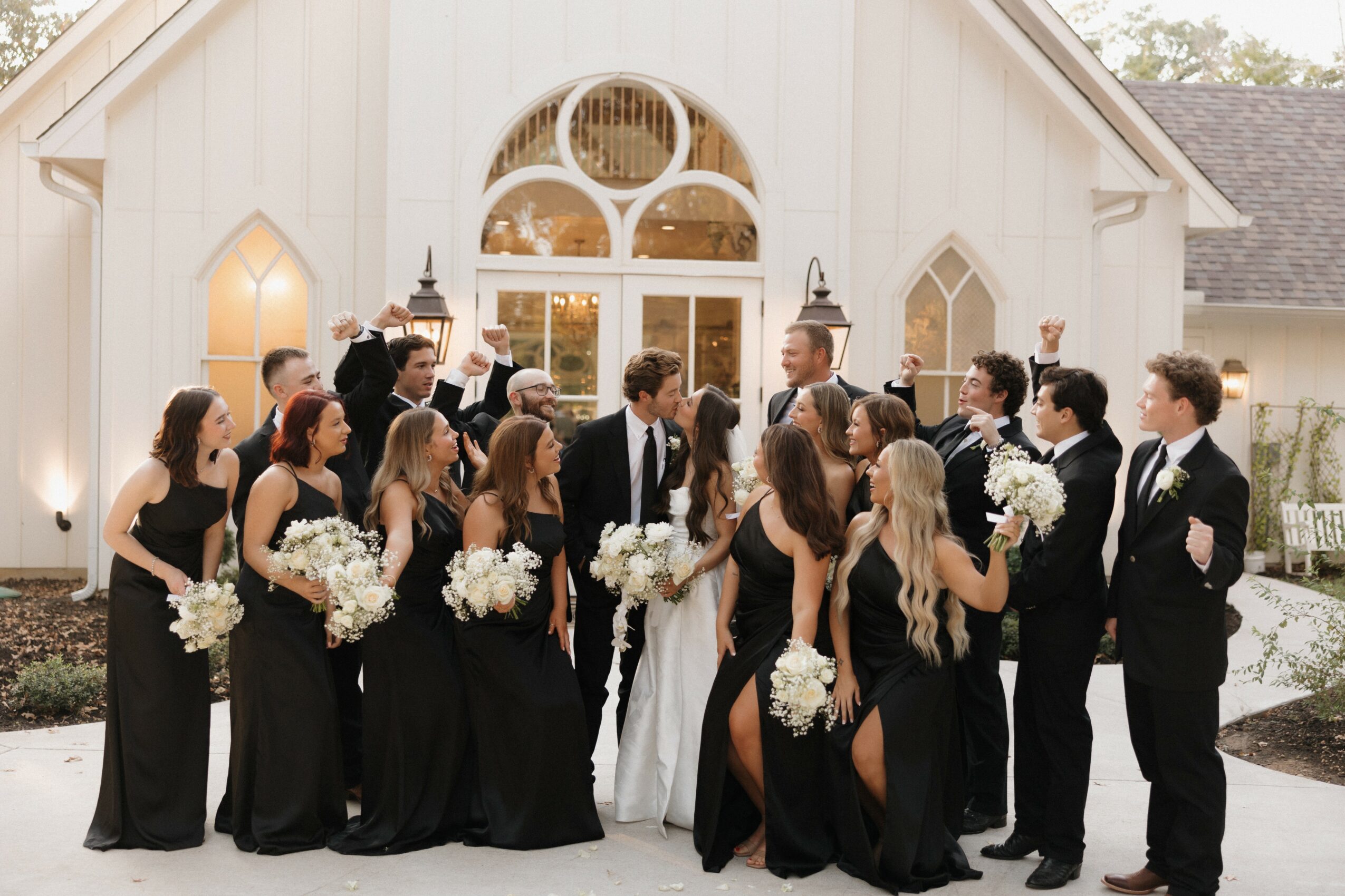 Group of people in formal attire standing outside a white building with arched windows, holding bouquets, celebrating a wedding.
