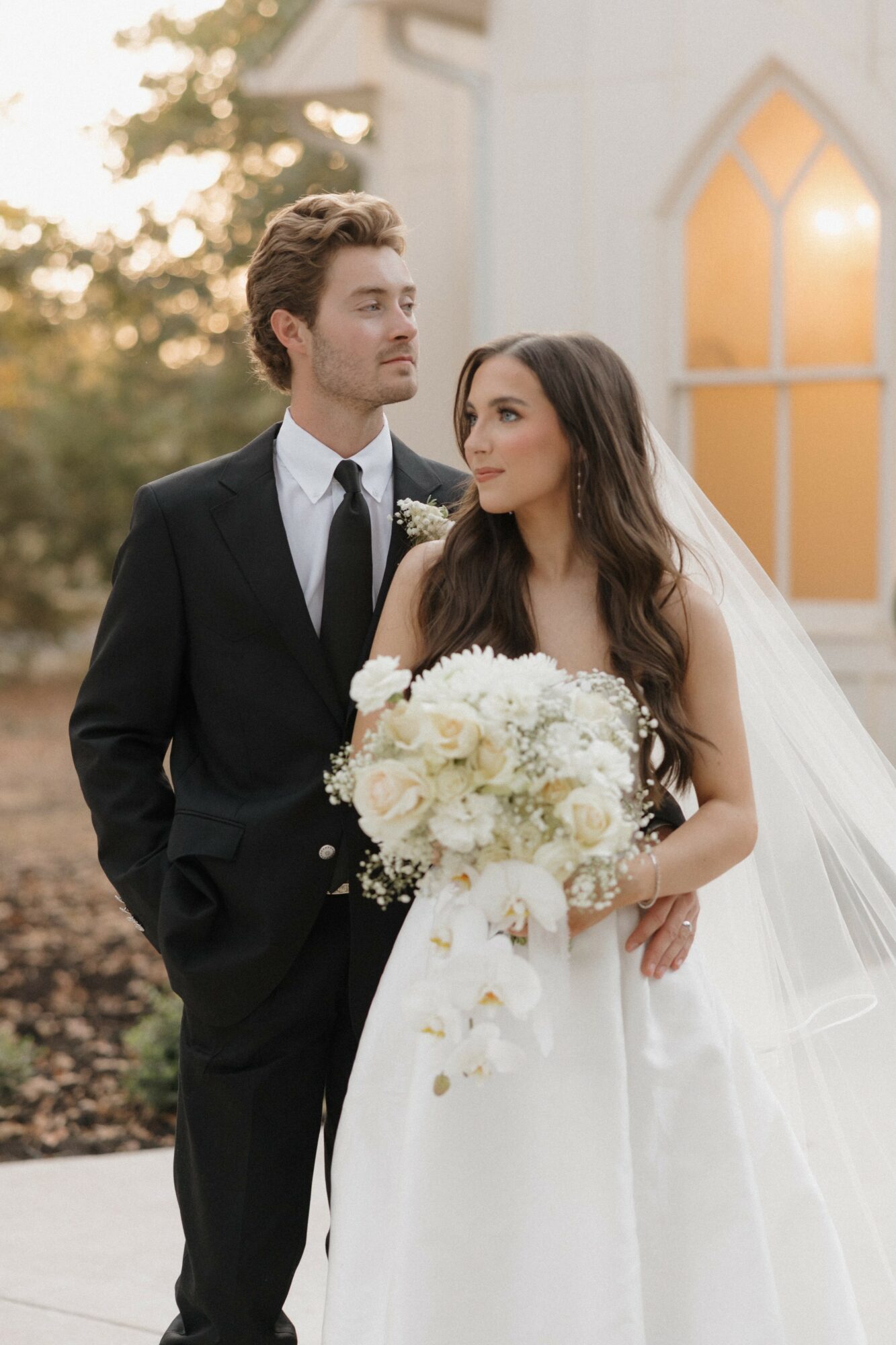 Bride holding a large bouquet of white flowers, standing beside groom outdoors near a building with arched window.