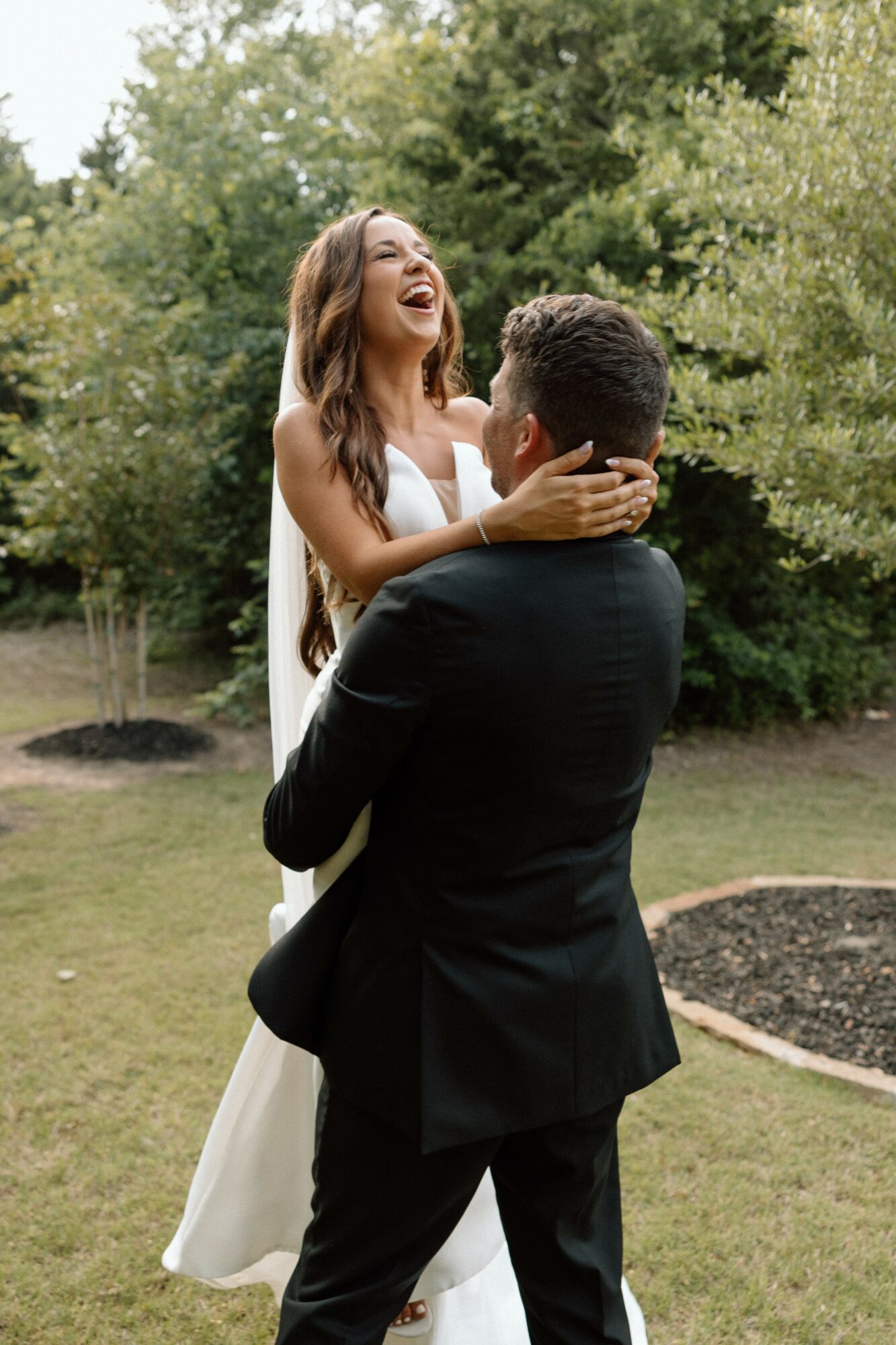A woman in a white dress is lifted by a man in a black suit outdoors, smiling and holding her face.