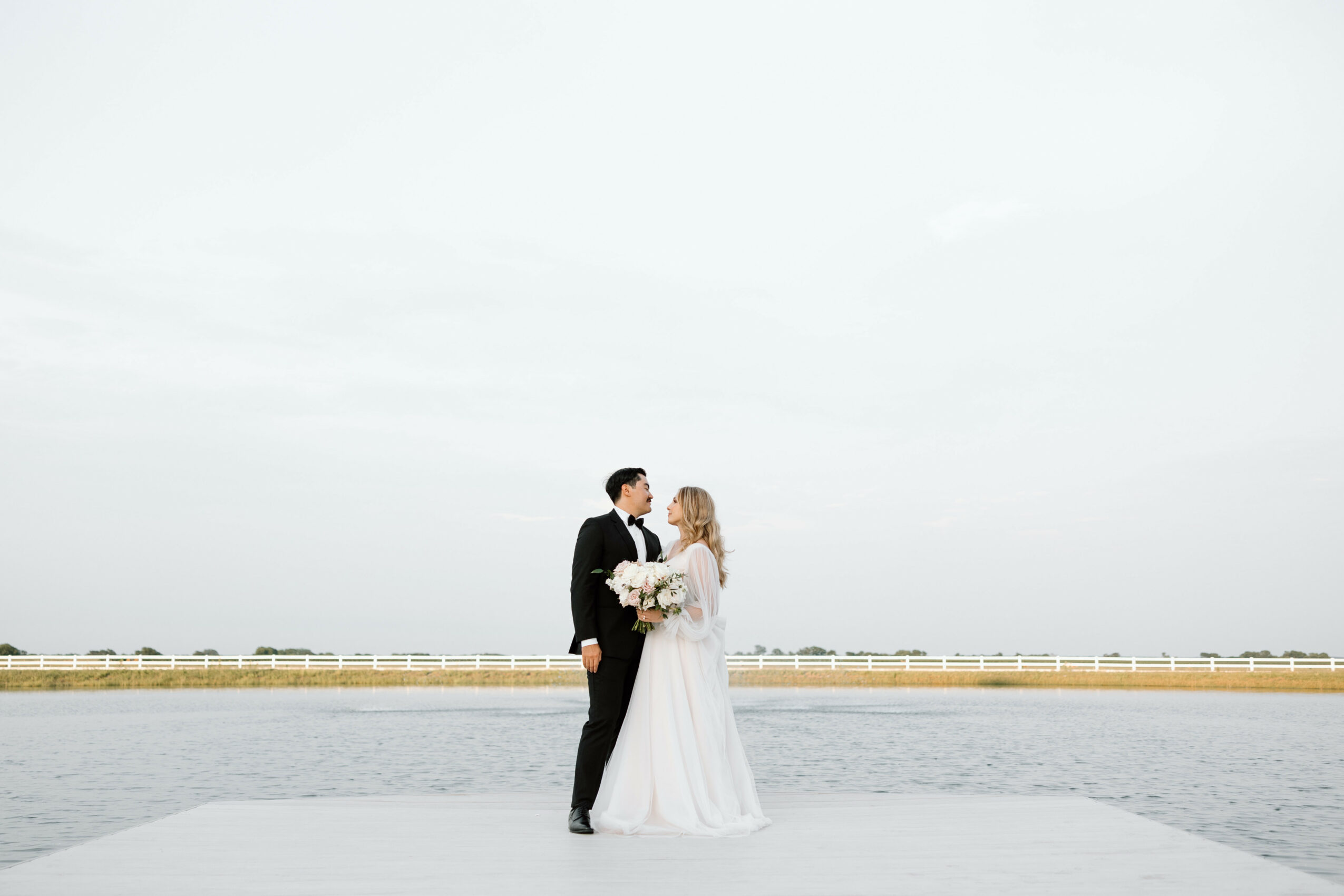 Bride and groom standing on a pier near water, holding flowers, facing each other, with a cloudy sky above.