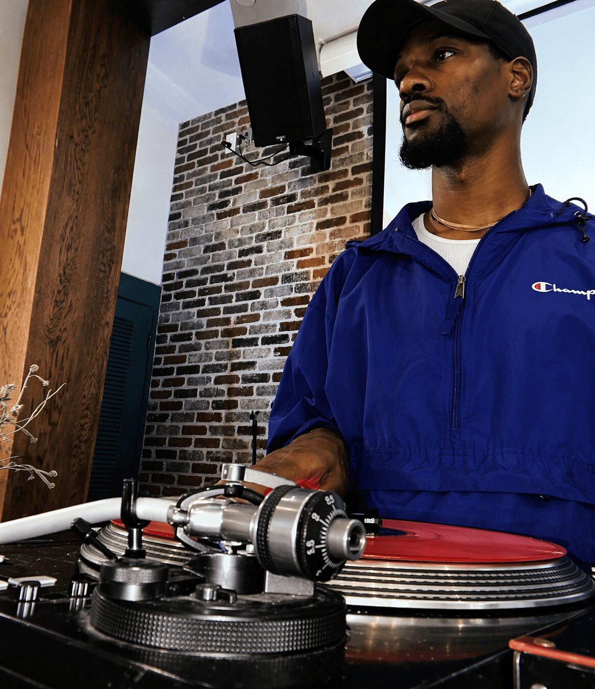 Man in blue jacket and black cap standing near turntable indoors with brick wall background.