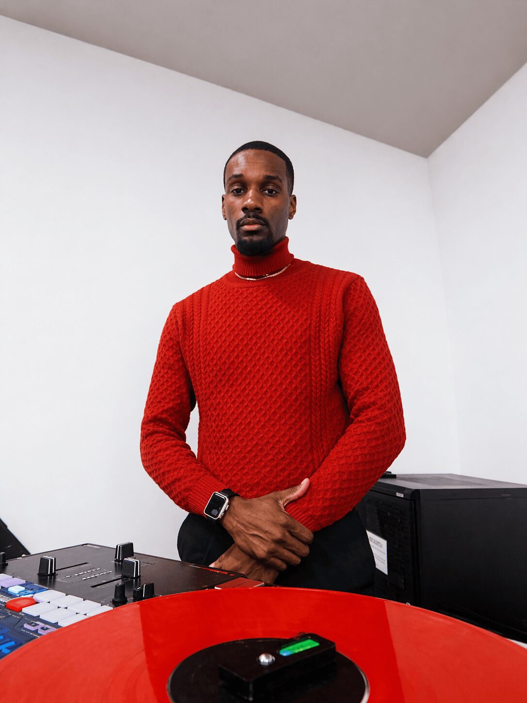 Man in red sweater standing with hands clasped in a room with white walls and a red table.