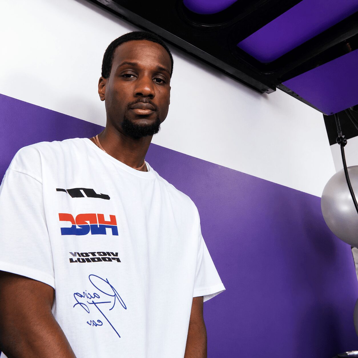 Young man with short hair and a beard wearing a white T-shirt with logos, standing indoors against a purple and white background.
