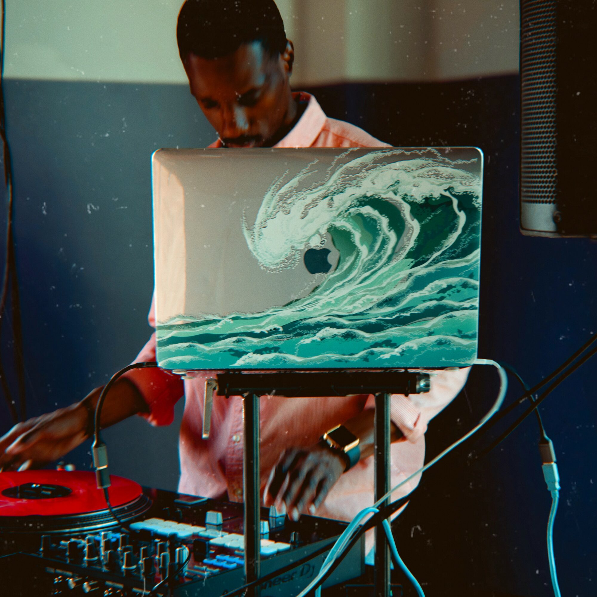 Person with dark skin and short hair using DJ equipment, laptop with ocean wave design, in a dimly lit room.