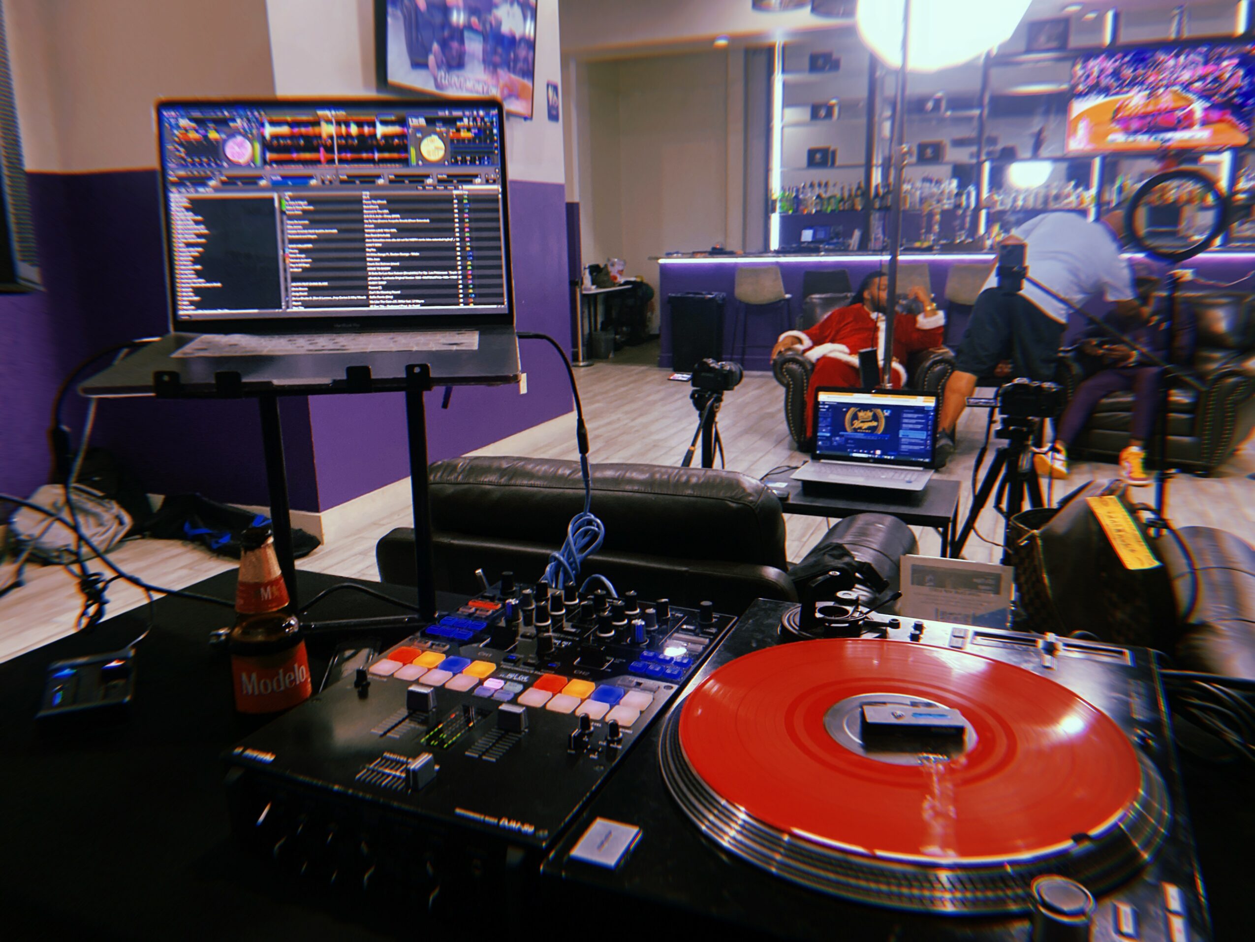 DJ equipment with a turntable, mixer, and laptop in a room with people and furniture in the background.