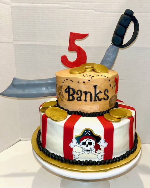 Two-tiered cake with pirate skull and crossbones, red and white stripes, gold coins, and a red number five topper.