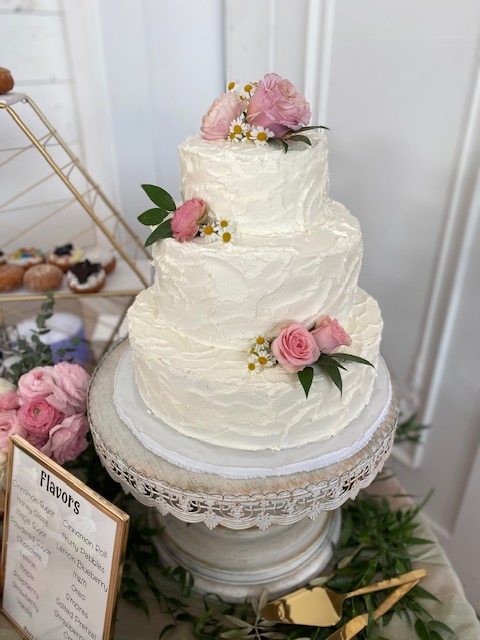 Three-tiered white cake decorated with pink roses and green leaves, displayed on a white ornate cake stand.