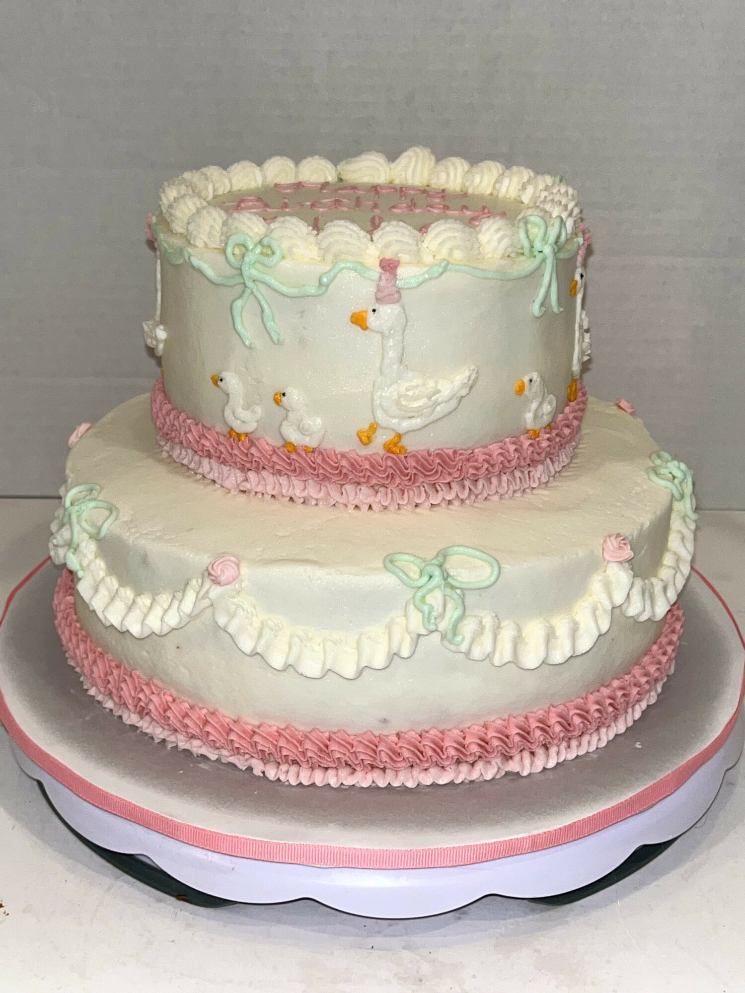 Two-tiered cake decorated with swans, ribbons, and pink accents, on a round cake board.