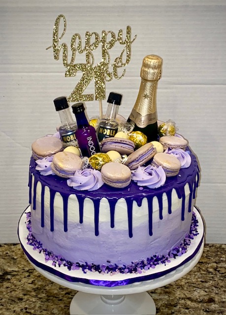 Celebration cake with purple and white icing, macarons, mini bottles, and a gold champagne bottle topper, with a 'happy 20' sign.