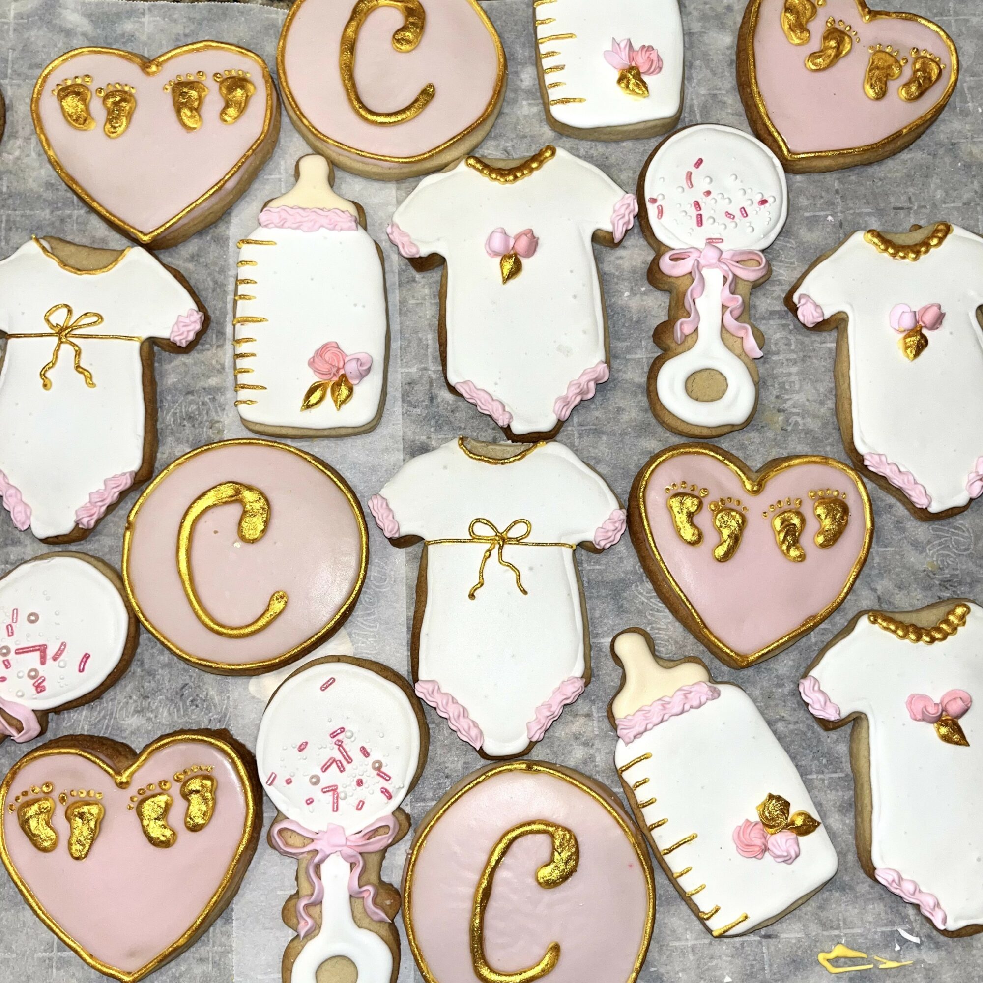Assorted baby-themed cookies with hearts, bottles, onesies, and a pacifier, decorated with pink, white, and gold icing.