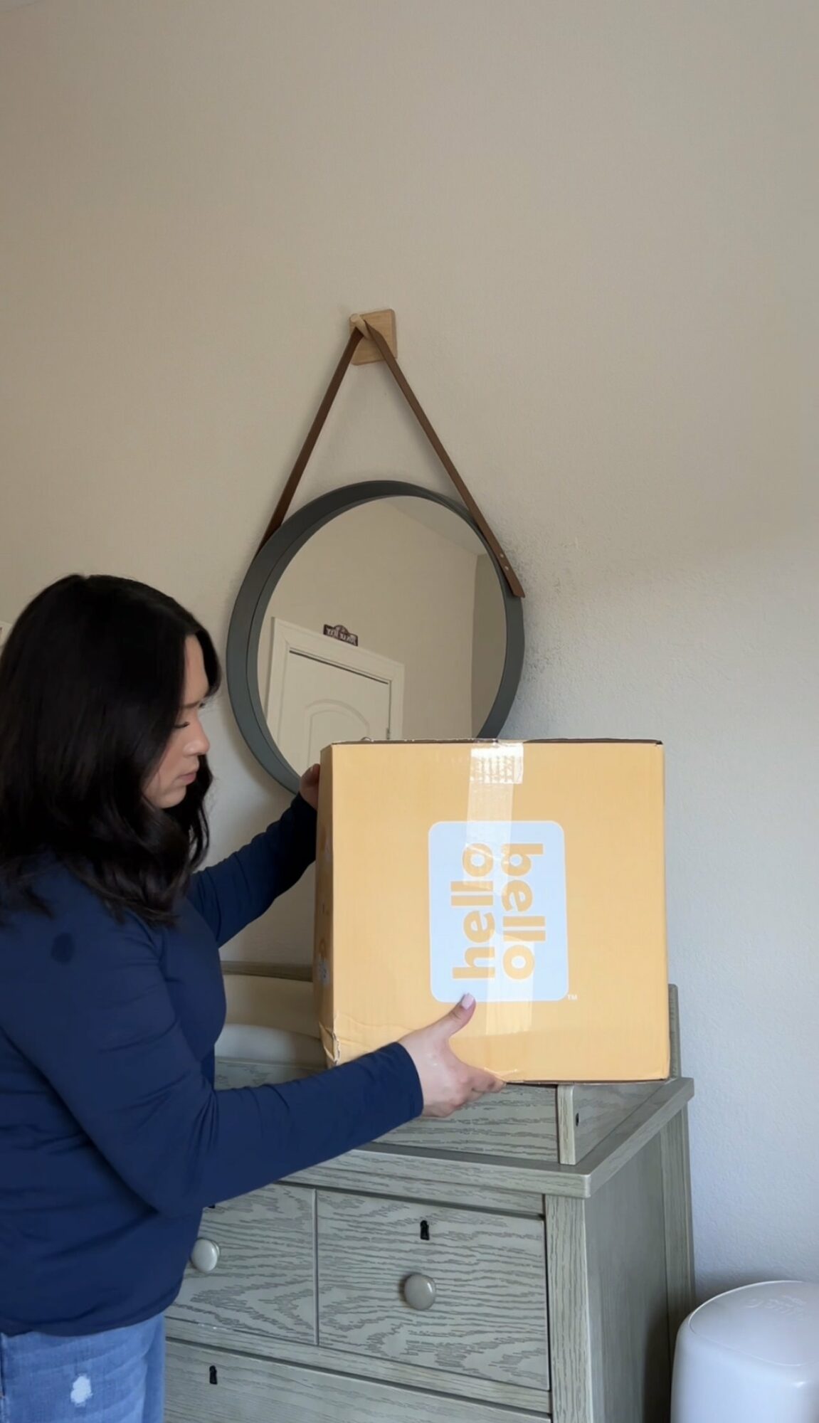 Woman with dark hair in a blue sweater places a box on a gray dresser, mirror on wall behind her.