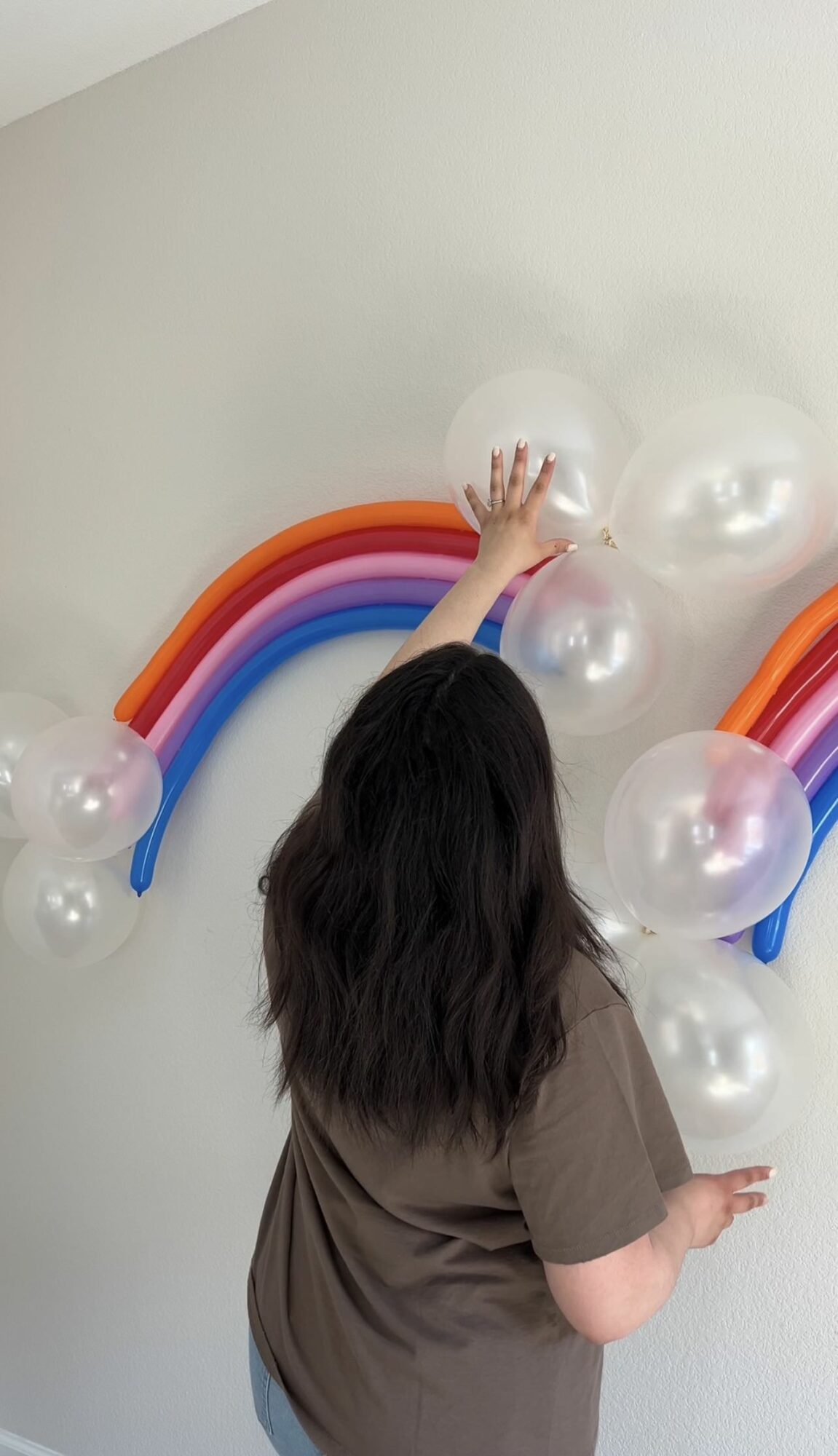 Person with long dark hair reaching for balloons and rainbow decorations on a wall.