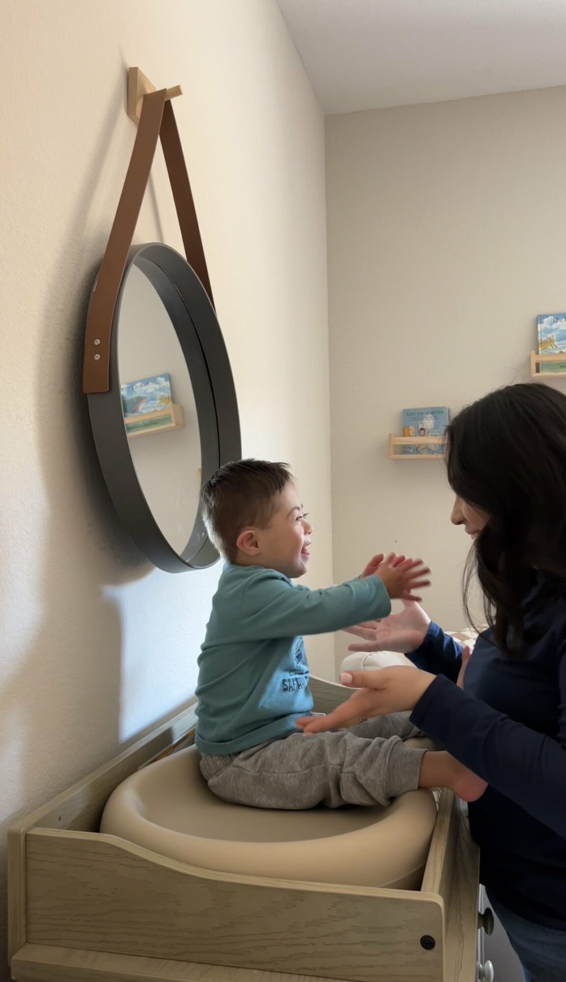 Child sitting on changing table, clapping hands with adult in a room with wall mirror and shelves.