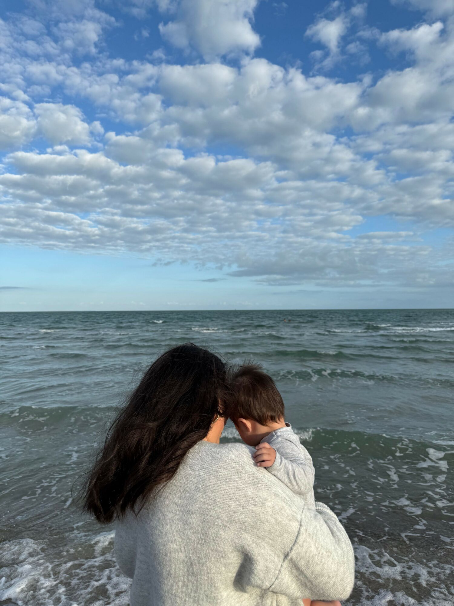 Person holding a child on a beach with ocean and cloudy sky in background.