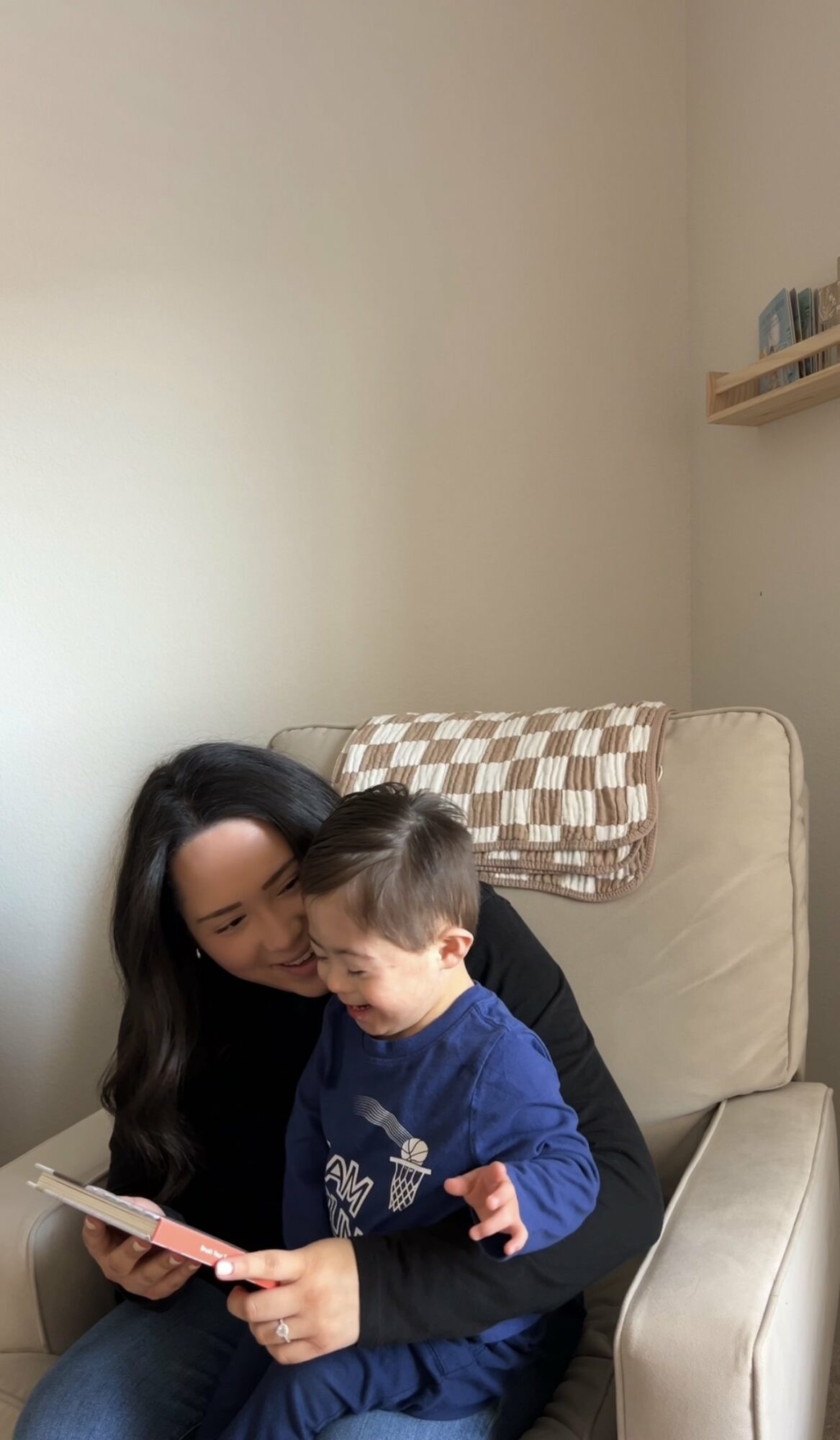 Woman and child sitting on a beige sofa, reading a book together, with a checkered pillow behind them.
