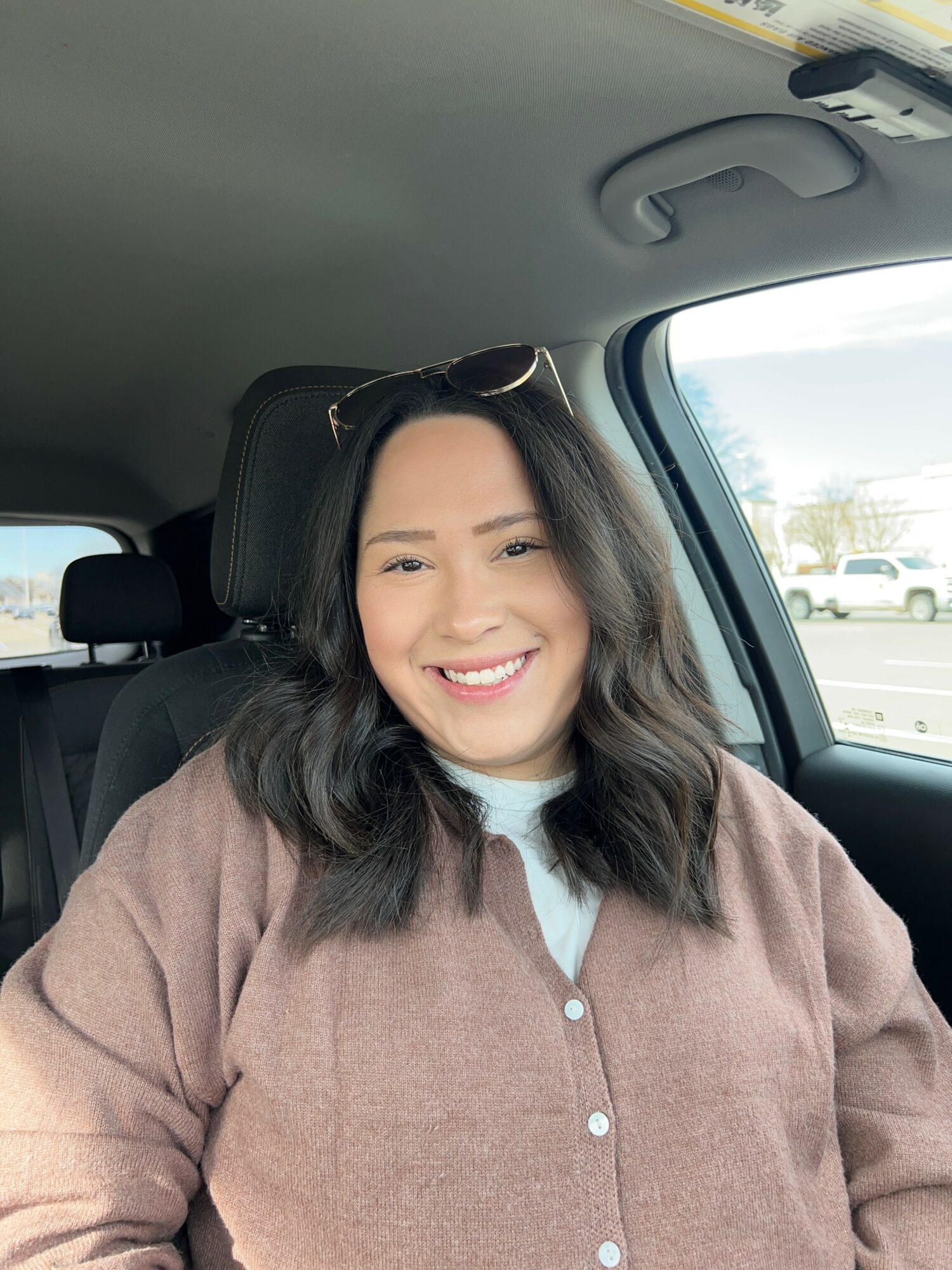 Smiling woman with wavy hair sitting in car, wearing a brown jacket and sunglasses on head.