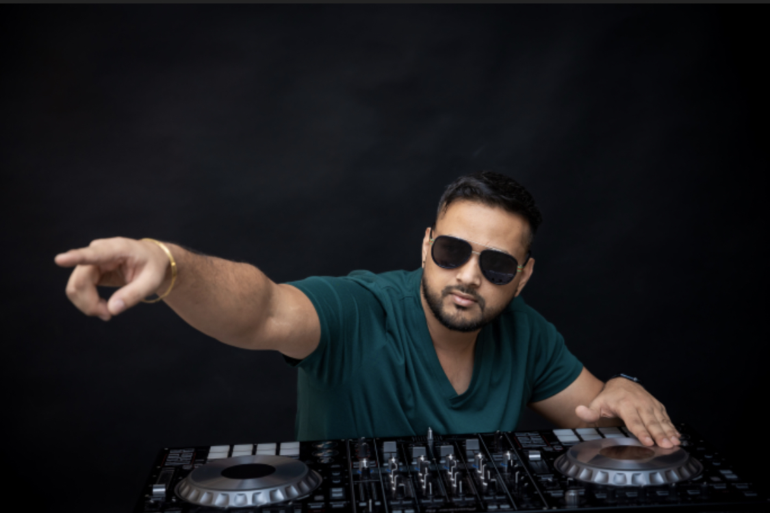 Man wearing sunglasses and a teal shirt points to the left while standing behind a DJ mixer.