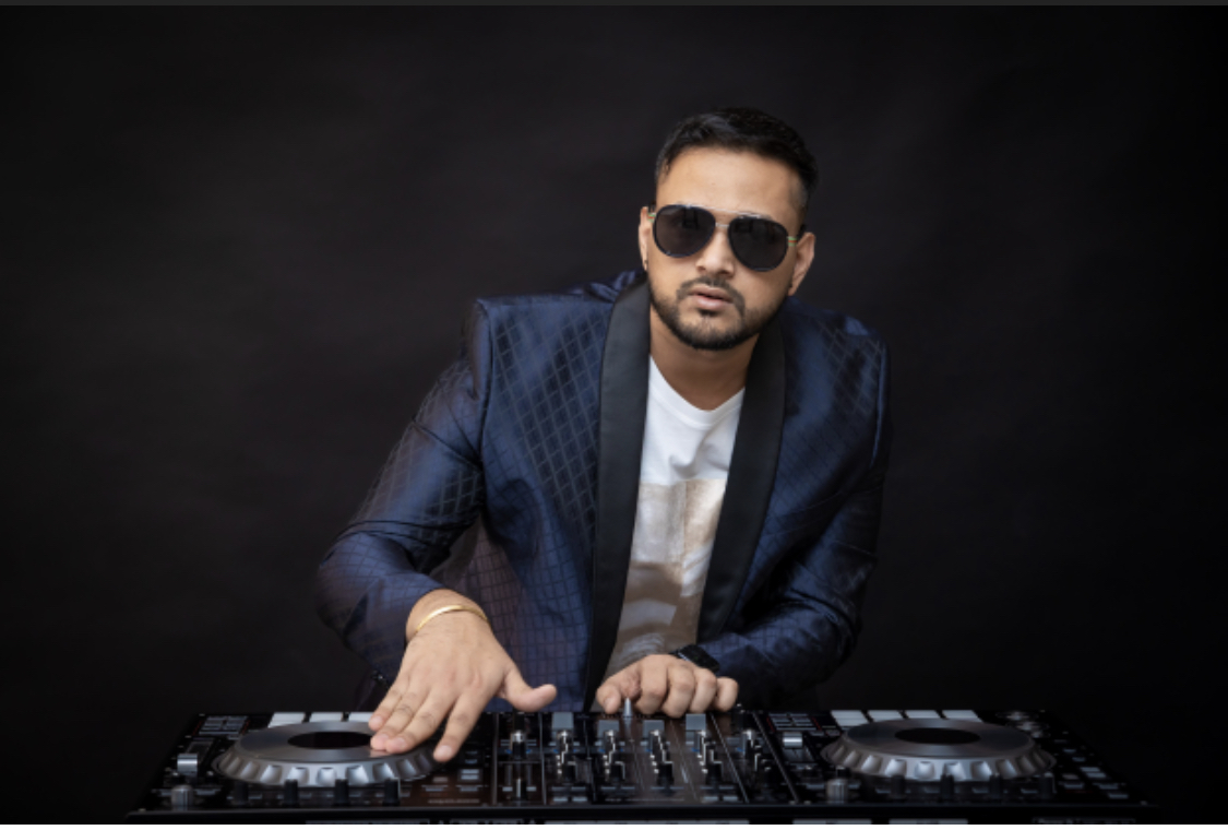 Man wearing sunglasses and a blue blazer, standing behind DJ equipment against a dark background.