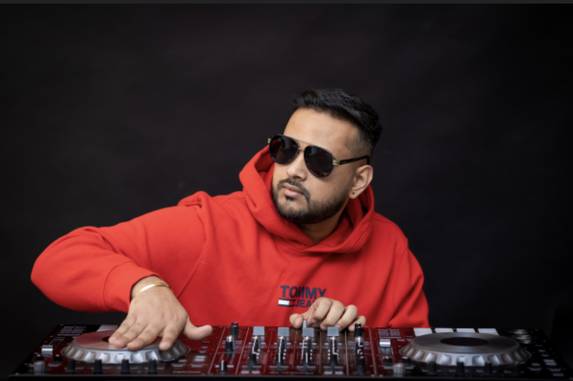 Man wearing sunglasses and a red hoodie operating a DJ mixer against a black background.