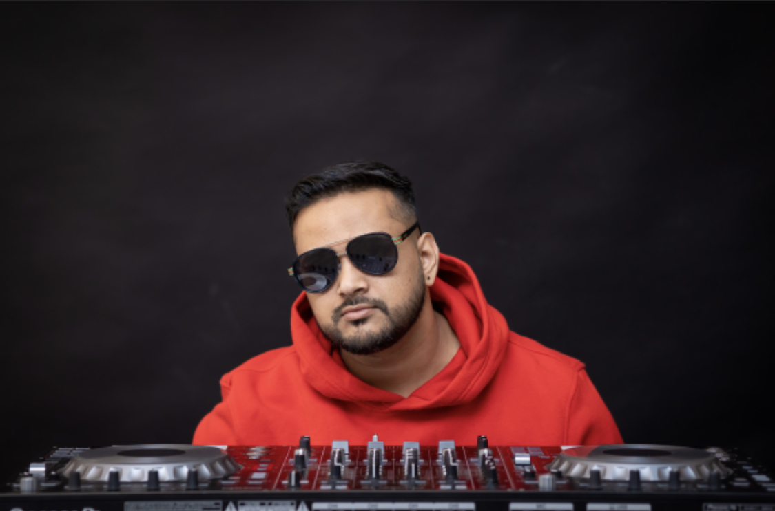 Man with sunglasses and a red hoodie stands behind DJ equipment against a dark background.