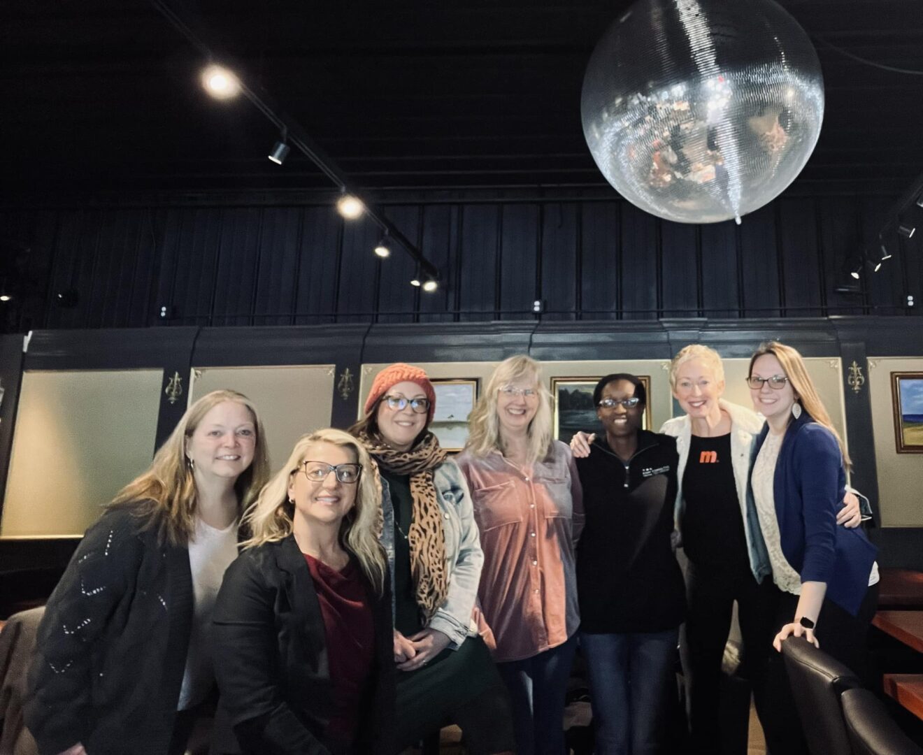 Group of seven women standing together indoors, with a large disco ball overhead and dark walls in the background.