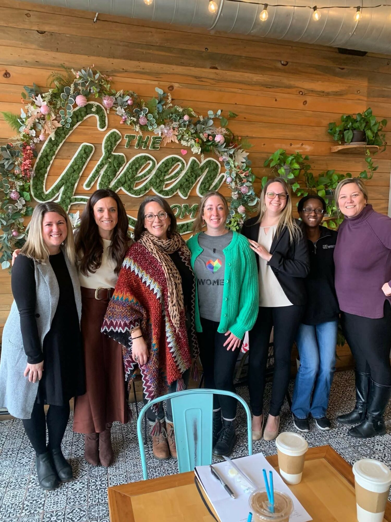Group of six women standing indoors in front of a wooden wall with a sign that says 'The Green'.