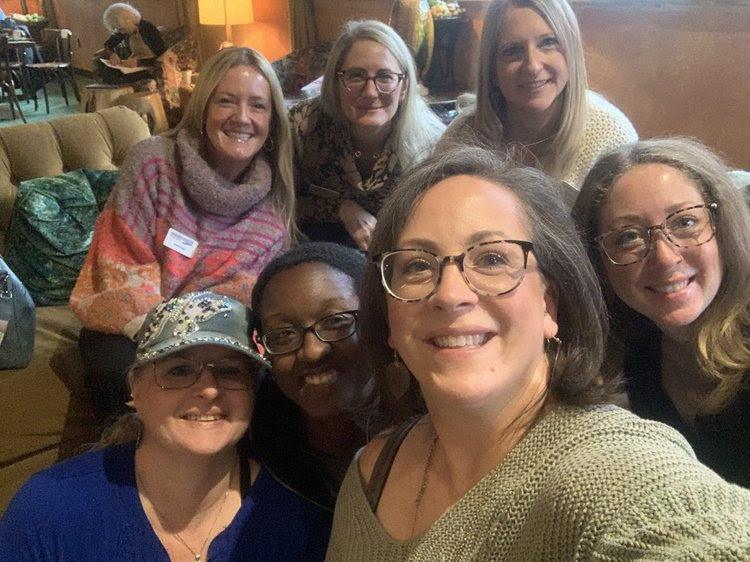 Seven women smiling together indoors, some wearing glasses and hats, in a cozy setting.