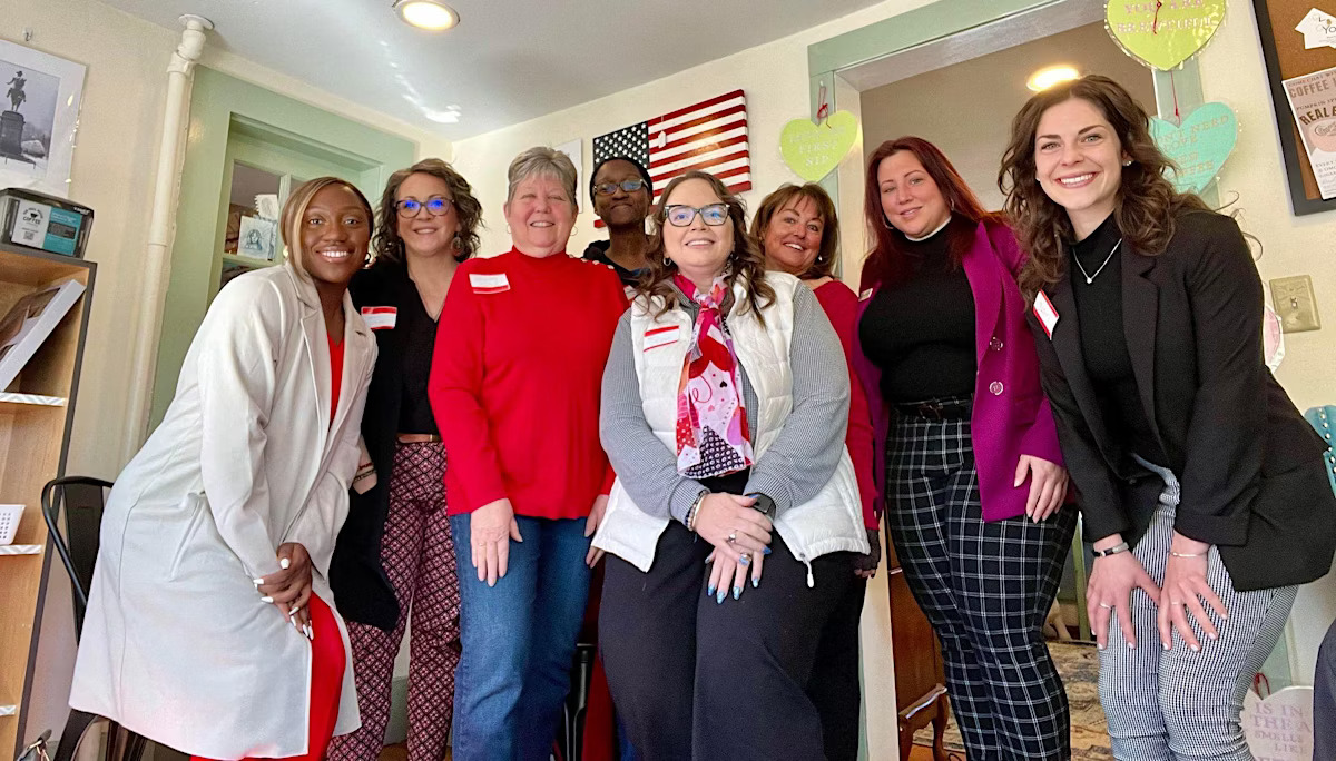Group of seven women standing together indoors, smiling, in a room with colorful decorations and American flag artwork.