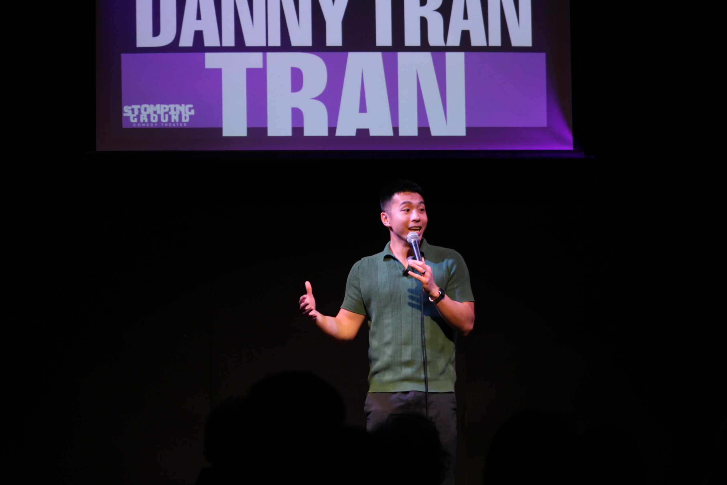 Person standing on stage holding a microphone, with a large purple and white sign above displaying text.