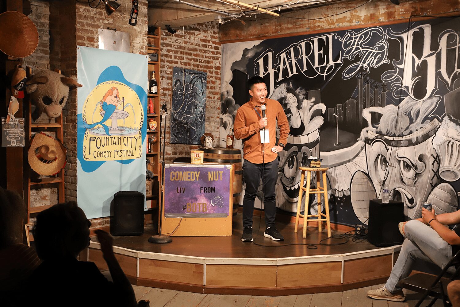 Person performing stand-up comedy on stage with microphone, in a venue with brick walls and artwork, audience seated.
