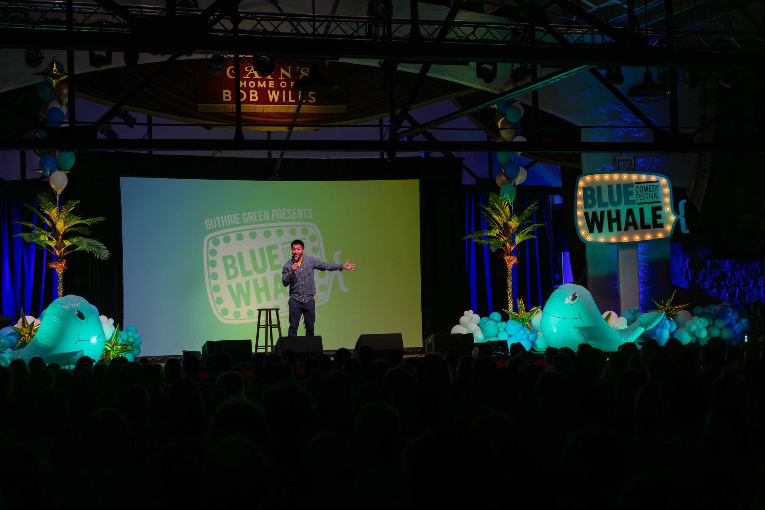 Person on stage with a microphone, large screen behind, decorated with blue whale and palm trees, audience in foreground.