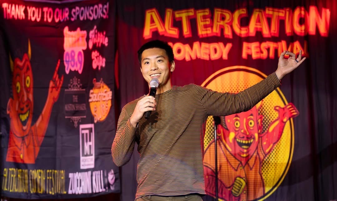 Male comedian on stage holding microphone, gesturing with hand, at Altercation Comedy Festival, colorful backdrop with cartoon character.