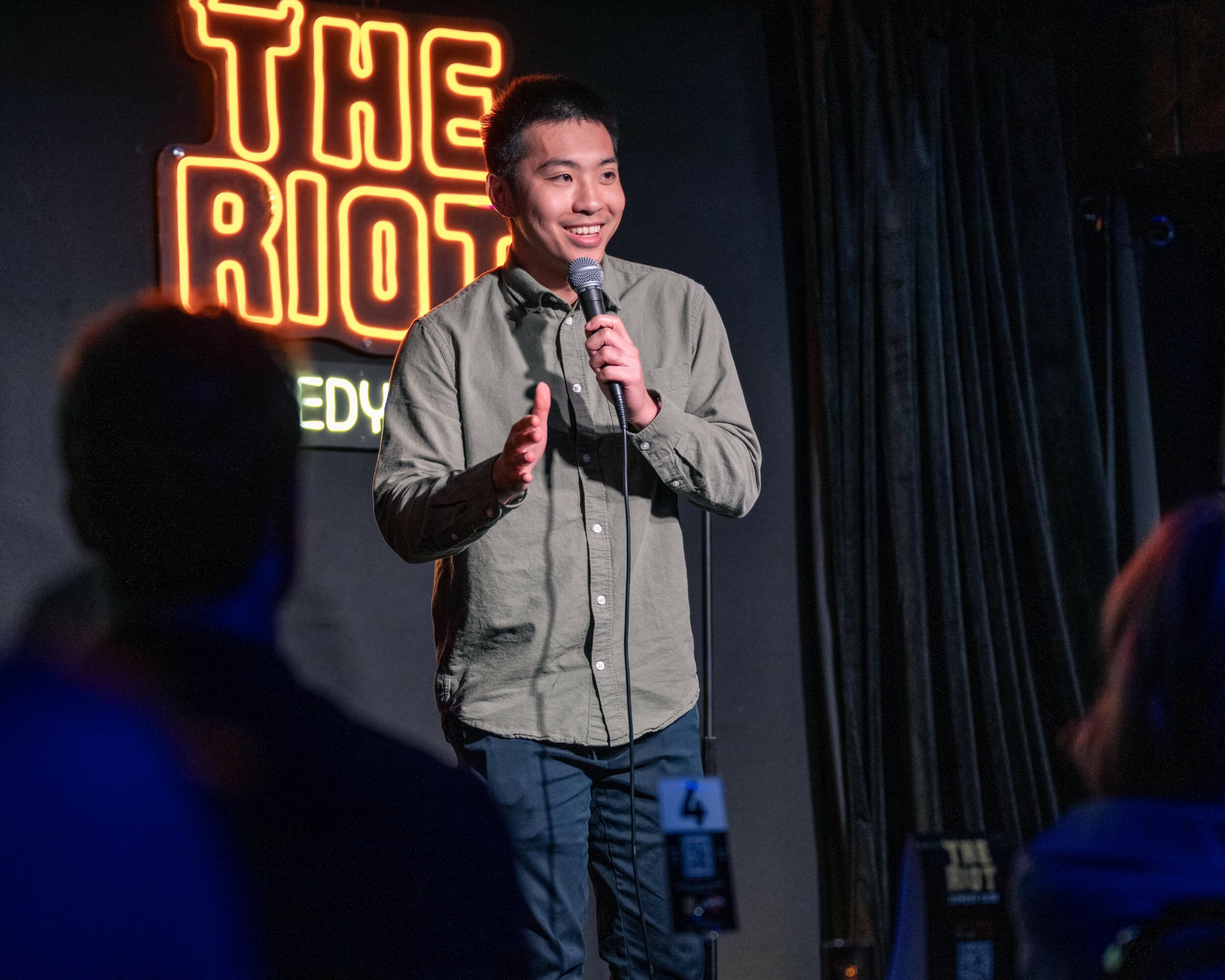 Person standing on stage holding a microphone, smiling, with a neon sign behind them that reads 'THE RIOT'.