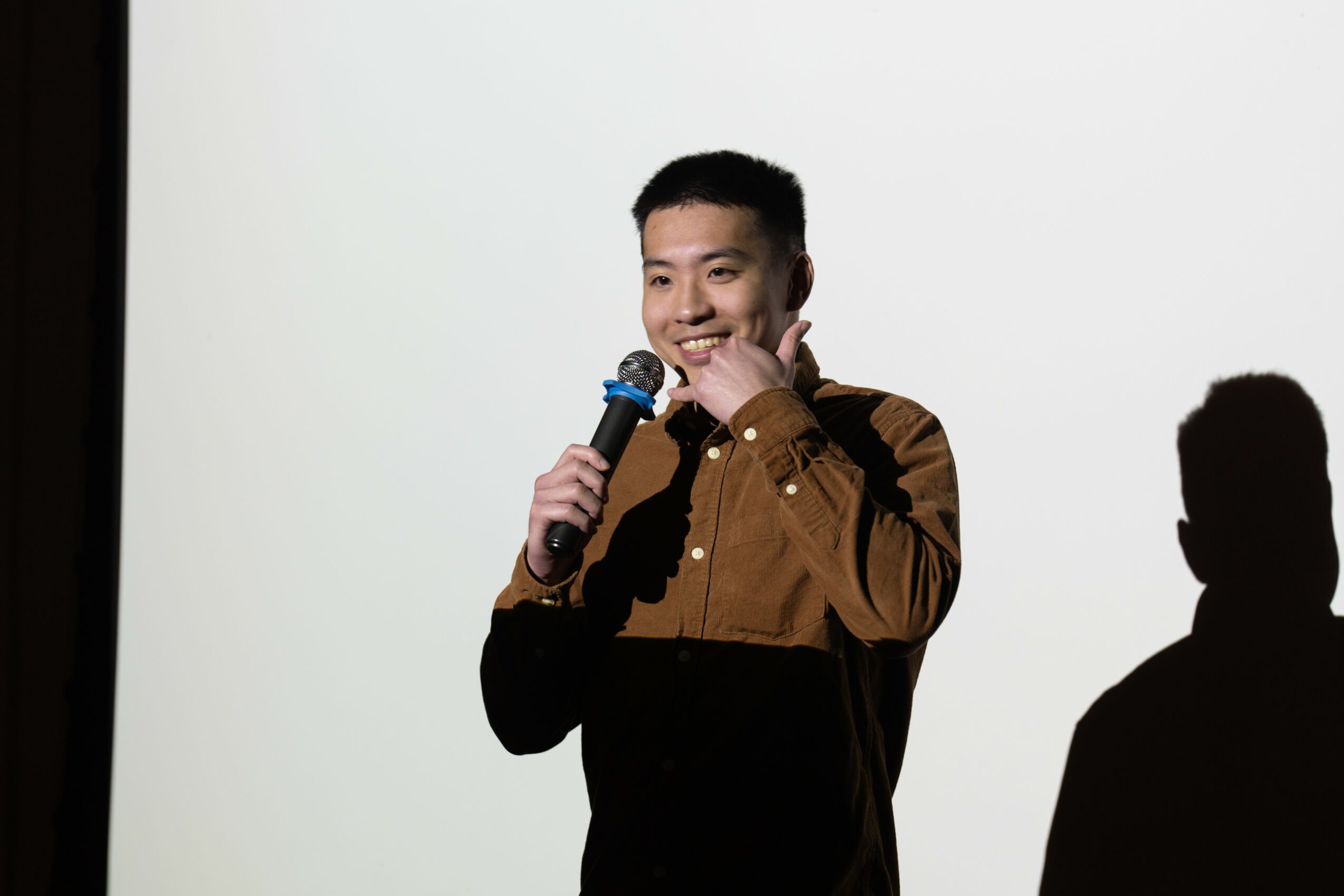 Young man holding a microphone, smiling, standing in front of a white background with a shadow cast on the wall.