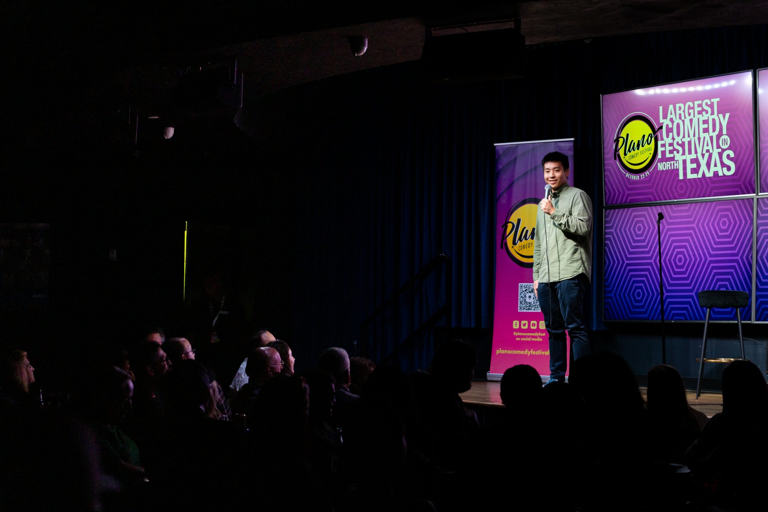 Person on stage speaking to audience with colorful background and banners, audience seated in darkened room.