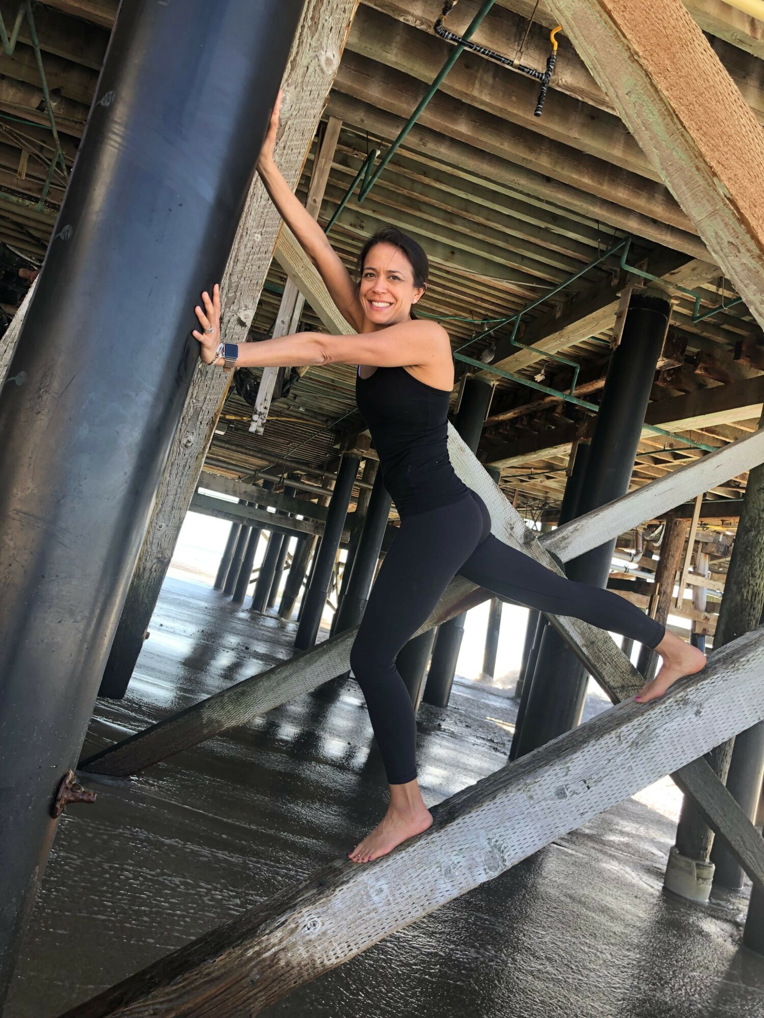 Woman in black outfit balancing on a wooden beam under a pier structure.