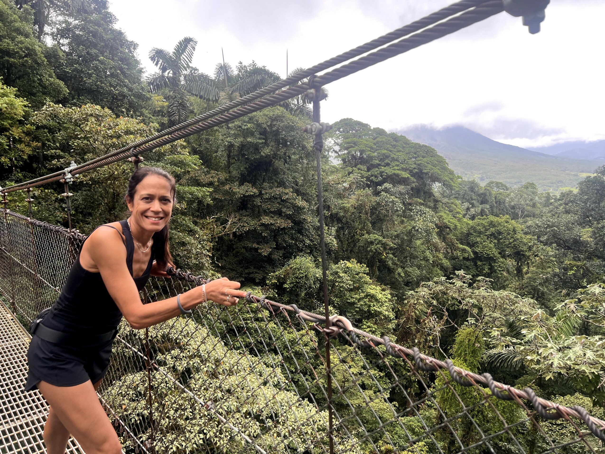 Woman smiling on a suspension bridge surrounded by lush green trees and mountains in the background.