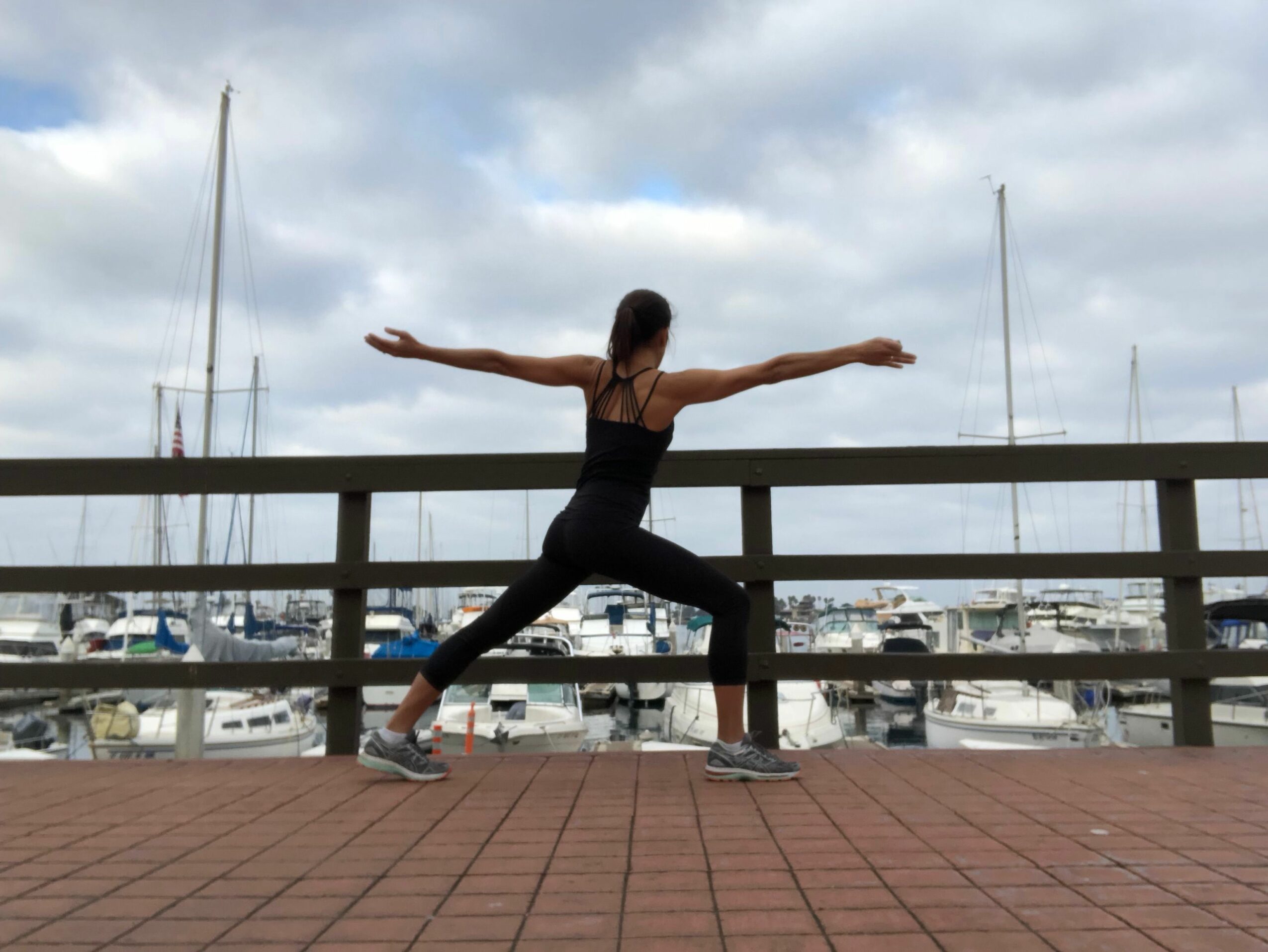 Person performing a star jump with arms and legs extended, standing on a brick surface near a marina with boats.