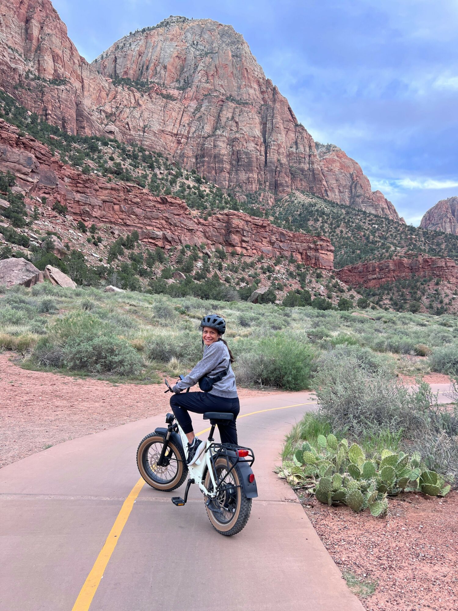 Person riding a bicycle on a paved trail with red rock formations and green vegetation in the background.